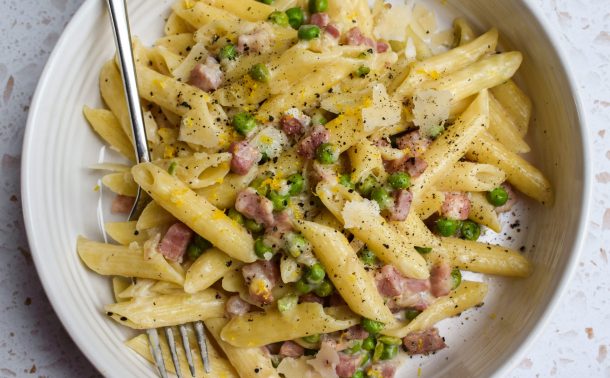 White bowl of pasta with pancetta and peas on the worktop.