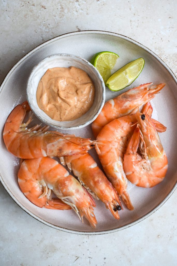 Pot of red curry mayonnaise on a plate of shell on prawns with lime wedges.